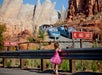 A young girl in a pink dress stands by a guardrail, watching cars on a race-themed amusement park ride, with red "been painted" and "he had just" signs nearby.