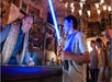 A boy holds a blue lightsaber while smiling at a man behind a counter in a themed, sci-fi setting, with other people watching in the background.