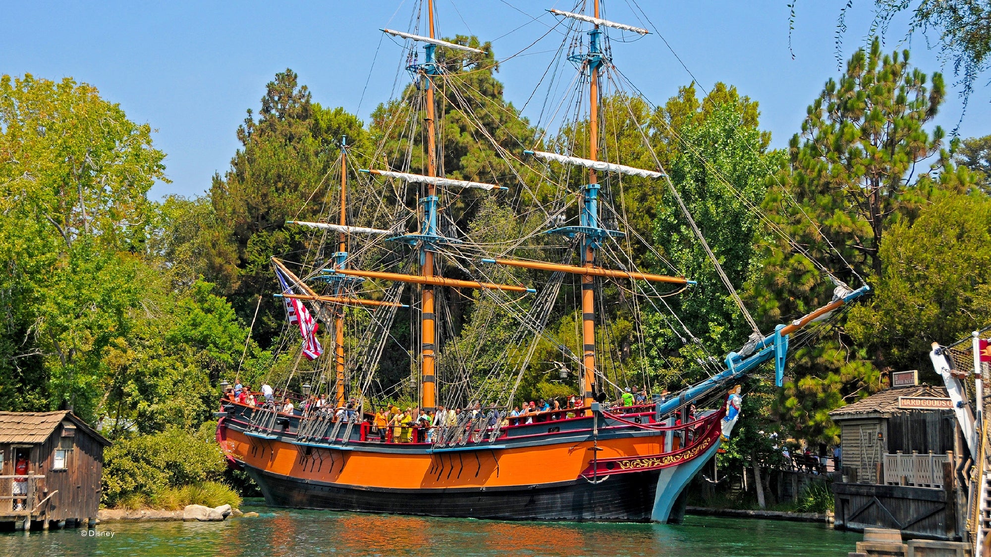 A large, colorful sailing ship with three masts carries passengers along a river surrounded by trees and wooden structures.