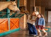A woman and a young girl stand smiling in front of horse stalls, looking at two horses inside a stable.