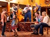 Two children dance in front of a seated couple while a bluegrass band plays on stage in a warmly lit, rustic venue.