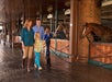 A family of four strolls through a horse stable, looking at the horses in their stalls. A woman and man walk with a young girl and boy, all dressed casually. The stable has a brick and wooden structure.