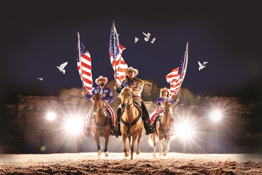 Three people on horseback carry American flags under bright lights with a dark sky and birds flying overhead.