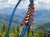 A roller coaster car with passengers goes through a vertical loop against a backdrop of forested mountains and a partly cloudy sky.