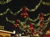 Large red bell decorations and green garlands with lights are strung overhead at night in an outdoor festive setting.