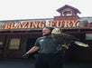 A man wearing gloves holds a bald eagle outside a building with a "BLAZING FURY" sign and a small fire station structure on the roof.