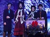 Four people in period costumes, three standing and one sitting in a sleigh, sing against a snowy backdrop, with the sleigh adorned with a red bow and greenery.