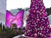 Large pink Christmas tree with metallic ornaments stands in front of a butterfly-shaped “Dolly” sign at an entrance, with people gathered around.