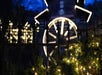 Holiday lights and greenery adorn a fence in front of a blurred, lit-up structure with wheel decorations against a dark evening sky.