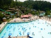 Aerial view of a crowded outdoor water park featuring a large wave pool with floating tubes, surrounding lounge chairs, multiple water slides, and a backdrop of trees.