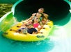 Four people ride a yellow inflatable raft down a green water slide at a water park, emerging into the sunlight.