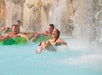People float on inner tubes along a water ride, passing under waterfalls, with one child in the foreground laughing and splashing.