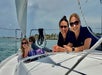 Two women and two children wearing sunglasses relax on the deck of a sailboat on a sunny day, with water and shoreline visible in the background.