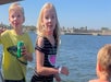 Three children are on a boat by the water, with one holding a can of Sprite; a shoreline with trees and buildings is visible in the background under a clear sky.