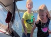 Two children on a boat; the boy is holding a can of Sprite and smiling, while the girl walks beside him. Water and shoreline are visible in the background.