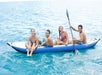 Four people sit together on a blue inflatable kayak in clear blue water, smiling and posing for the photo. One person holds a paddle.