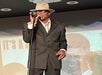 A man in a suit and hat sings into a microphone on stage, standing in front of a projected image and text.