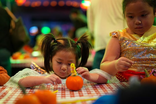 Two young girls decorate small pumpkins at a table covered with a red and white checkered cloth in a festive, colorful indoor setting.