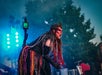 A performer in dramatic costume and makeup shouts on an outdoor stage, holding a green prop with trees and stage lights in the background.