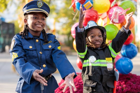 Two children in police officer and firefighter costumes smile and raise their arms in front of a display of colorful balloons.