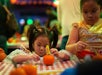 Two young children sit at a table, decorating small pumpkins with craft materials at a festive indoor event.