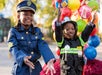 Two children dressed as a police officer and firefighter smile and raise their hands, with colorful balloons in the background.