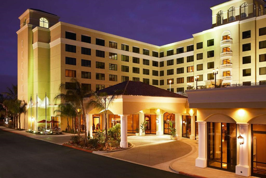 A well-lit hotel building at dusk with multiple floors, a curved driveway, and entrance columns. The sky is a deep blue.