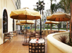 Outdoor restaurant patio with brown tables and chairs under beige umbrellas, next to a yellow building.