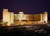 Night view of a large, well-lit hotel with several floors, surrounded by palm trees and a parking lot filled with cars.