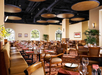 Restaurant interior with wooden tables, cushioned chairs, and a patterned carpet. Arched windows allow natural light, and ceiling features circular panels. Tables are set for dining.