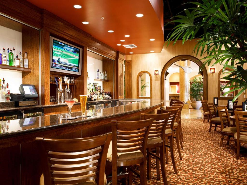 A dimly lit bar area with a long countertop, wooden chairs, a television mounted on the wall, and a variety of drinks displayed behind the counter.