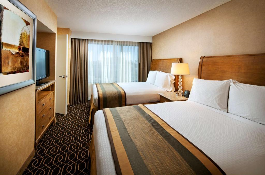 Hotel room with two double beds, a nightstand with a lamp, a flat-screen TV, a dresser, and a window with closed curtains. The walls are beige, and the carpet has a geometric pattern.