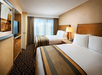 Hotel room with two double beds, a nightstand with a lamp, a flat-screen TV, a dresser, and a window with closed curtains. The walls are beige, and the carpet has a geometric pattern.