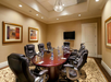 A conference room with a polished wooden table surrounded by nine leather chairs, water bottles, pens, and a mounted TV on the wall. Two framed abstract paintings decorate the cream-colored walls.