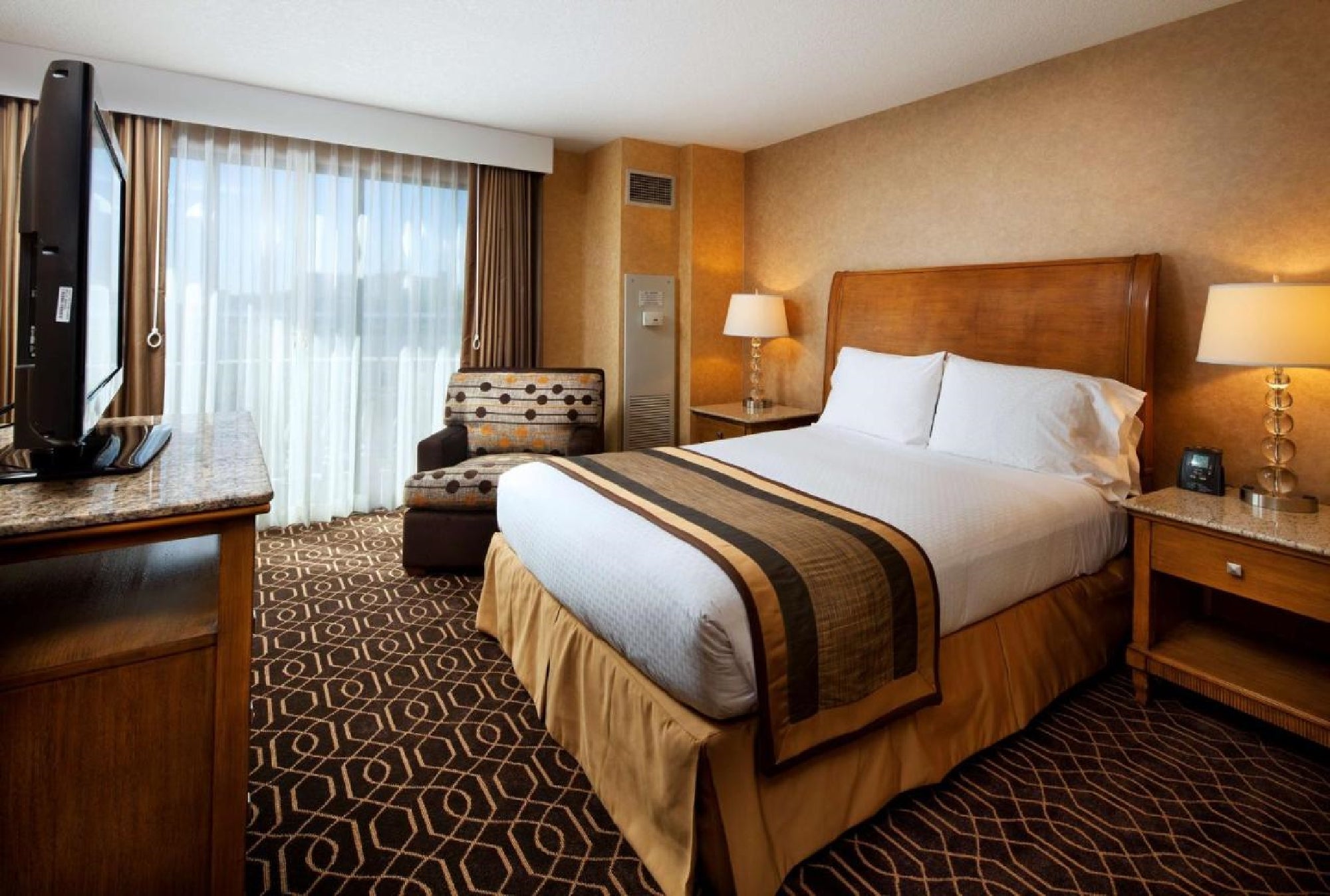 A hotel room with a neatly made bed, two bedside tables with lamps, a patterned chair, a TV on a stand, and a window with sheer curtains.