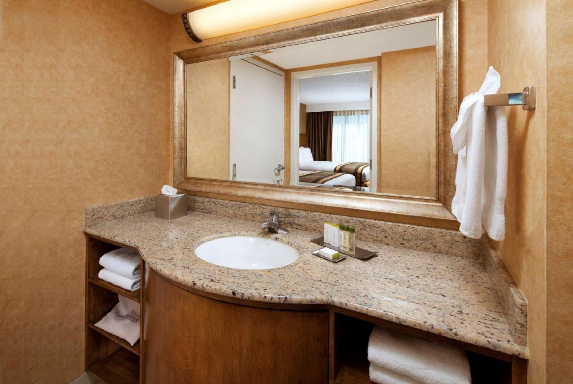 A hotel bathroom with a granite countertop, sink, mirror, and towels. A bed and window are reflected in the mirror.