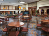 Spacious hotel lobby with circular wooden tables and chairs, patterned carpet, and warm lighting. Stone accent walls and greenery in the background.