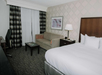 Hotel room with a bed, sofa, small table, and desk with chair. A lamp is on the nightstand, and artwork hangs on the patterned wall. Curtains cover a large window or sliding door.