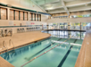Indoor swimming pool with lanes, surrounded by pool chairs and large windows. Ceiling lights illuminate the area.