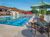 Outdoor swimming pool with lanes, surrounded by lounge chairs and tables with umbrellas, next to a building.