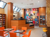Modern cafe interior with orange chairs, wooden tables, and a patterned wall. Shelves display various products, and a counter with a display case is on the left.