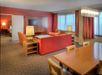 A hotel suite with a living area featuring red sofas, a wooden coffee table, a TV on an accent wall, and a dining table with chairs. A bedroom is visible through an open door.