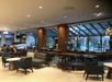 A modern hotel lobby with seating areas, tables, and large windows. The space features overhead lighting and decorative plants.