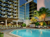 Outdoor hotel pool area with lounge chairs, umbrellas, and palm trees, situated next to a building with reflective windows.