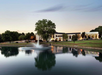 A large building with illuminated windows is set behind a tranquil pond with a fountain, surrounded by trees and grass, under a dusk sky.
