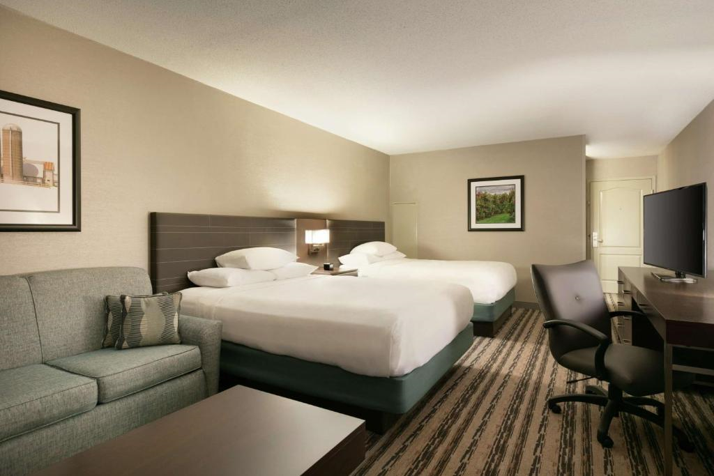 Hotel room with two double beds, a sofa, and a desk with a chair. The walls are decorated with framed art. A television is placed on a dresser.