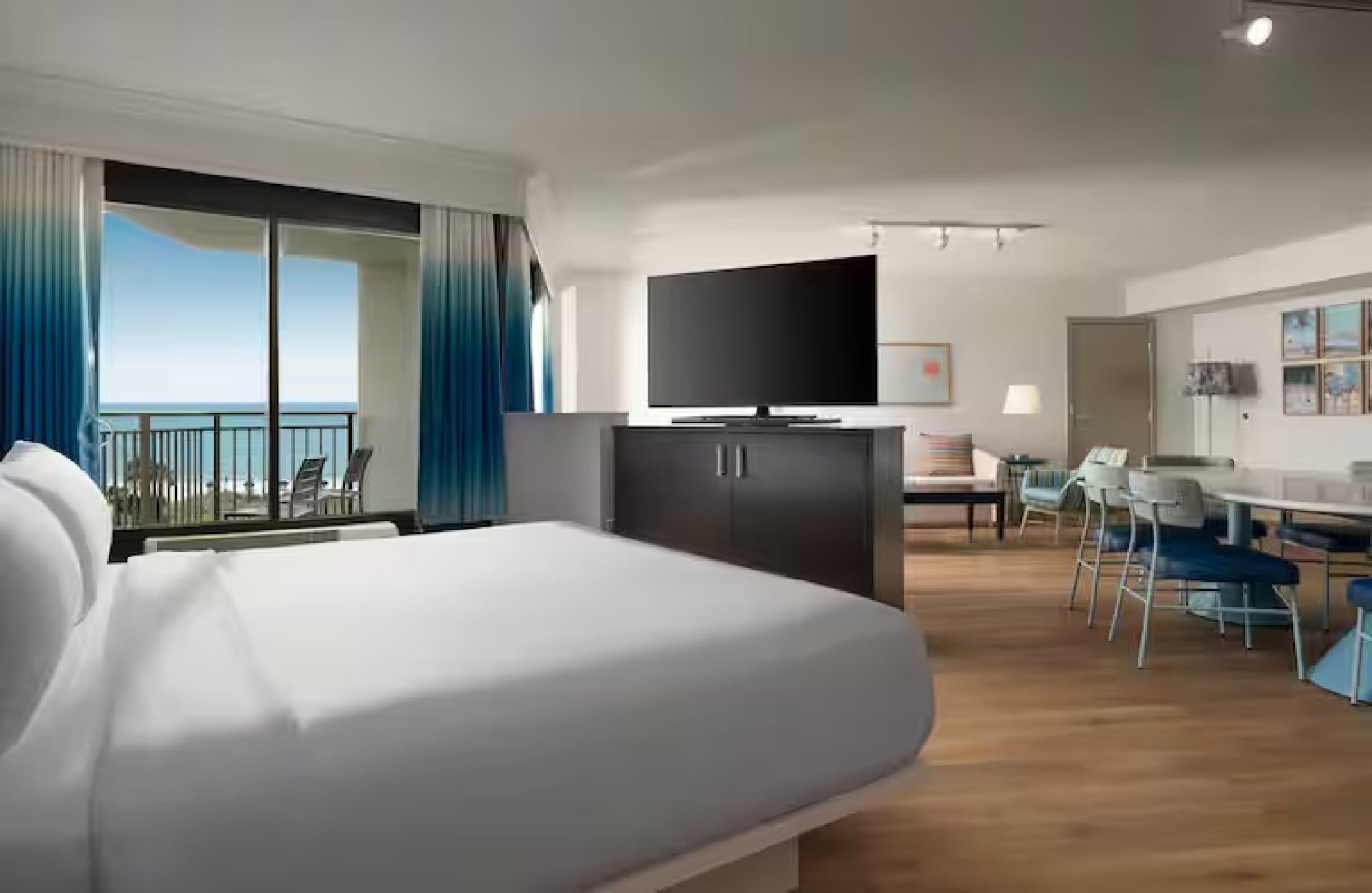 Modern hotel room with a large bed, flat-screen TV, seating area, and dining table. A balcony offers a view of the ocean, with blue curtains framing the sliding glass door.