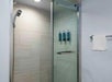 Glass-enclosed shower with beige tiles, wall-mounted showerhead, and three soap dispensers. A towel hangs on a rack outside the shower.