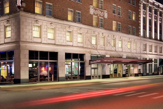 Street view of a DoubleTree Hotel by Hilton, featuring a brick and stone facade with decorative carvings and a red canopy over the entrance. Light trails from passing vehicles are visible.