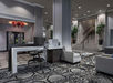 Modern hotel lobby with patterned carpet, a desk with a computer, two gray chairs, a glass coffee table, and an ornate red chandelier. A staircase with potted plants is in the background.